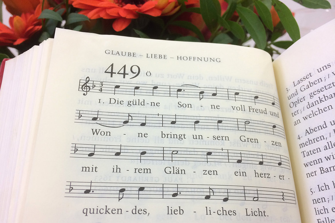 Seite aus dem Kirchenliederbuch, Lied 449: Die güldene Sonne, mit Noten, oberhalb des Buches sind ansatzweise rote Blumen mit grünen Blättern zu sehen