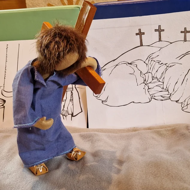 Eine kleine Holfigur mit blauem Gewand trägt ein braunes Holzkreuz auf der linken Schulter - Im Hintergrund ein Bild einer Fels-Erhöhung und darauf drei Kreuze