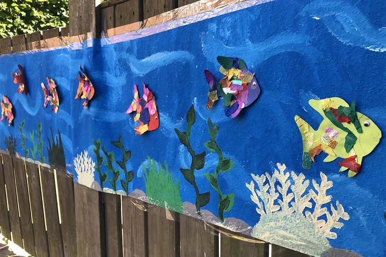 Aus Papier gebastelte bunte Fische auf einem langen blauen Papierstreifen angebracht, welches mit Wasserpflanzen bemalt ist. Der Streifen ist an einem braunen Lattenzaun befestigt.