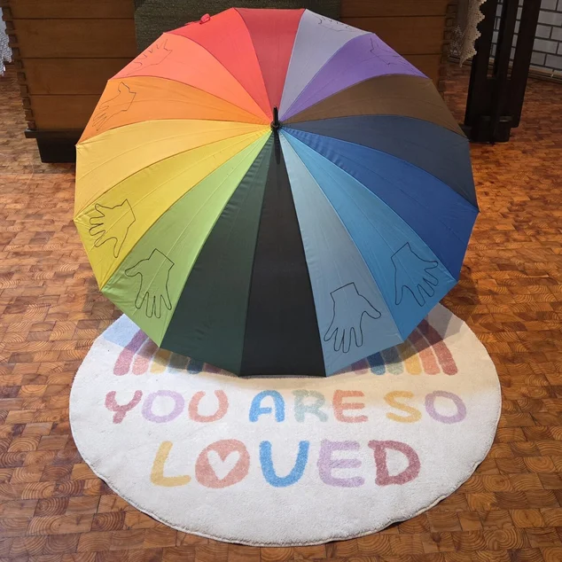Ein gegenbogenfarbener Regenschirm aufgespannt über einem runden weißem Kreis mit bunter Aufschrift "YOU ARE SO LOVED"